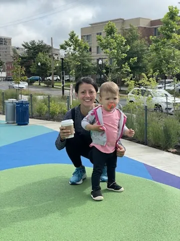 Marie holding Zoe at a colorful playground with coffee in hand