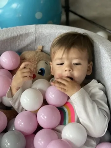 Zoe sitting in a soft bin filled with pink and white balls while holding a teddy bear