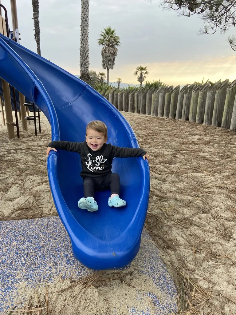 Zoe laughing at the bottom of a blue playground slide near the beach