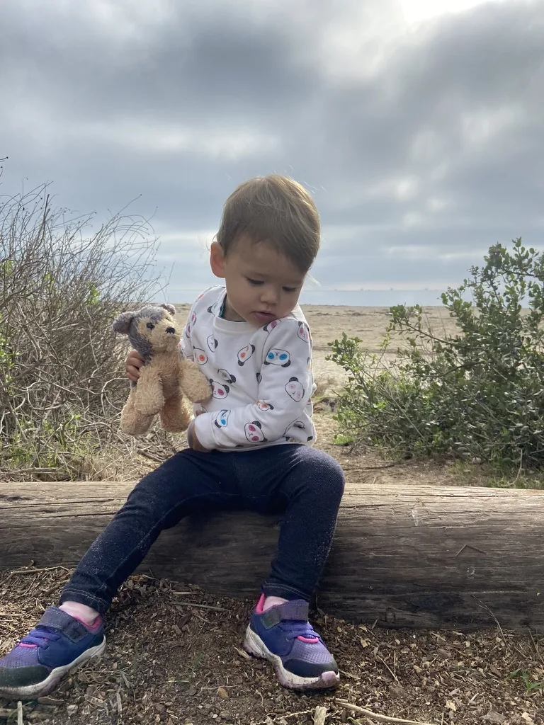 Zoe sitting on a beach log with a small stuffed bear
