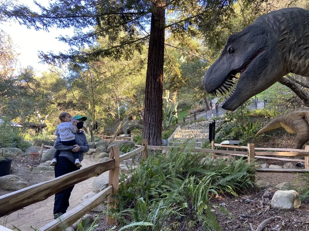 Marie holding Zoe near a dinosaur sculpture at a museum trail