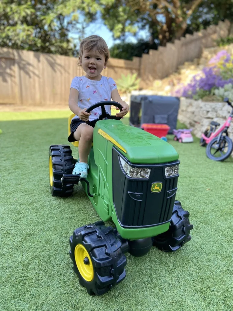 Zoe riding a toy John Deere tractor in a backyard