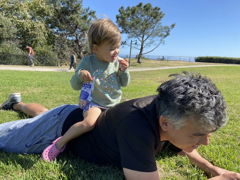 Zoe sitting on Andrew's back in the grass while holding a yogurt pouch