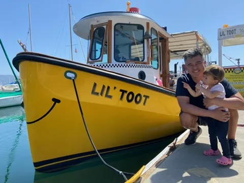 Andrew crouching with Zoe beside the yellow Lil' Toot boat