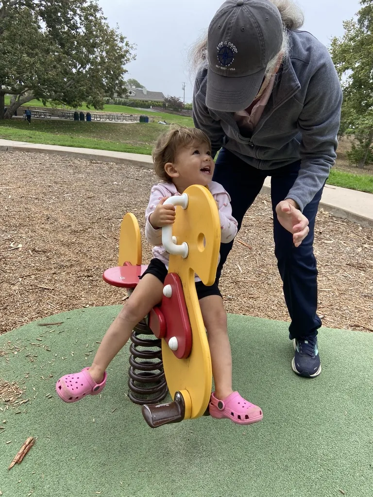Zoe riding a yellow playground spring toy while a family member leans toward her